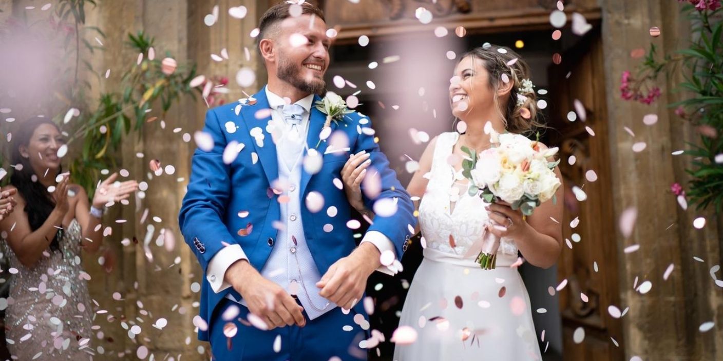 Mariés souriants sous une pluie de confettis roses et blancs à la sortie de leur cérémonie de mariage